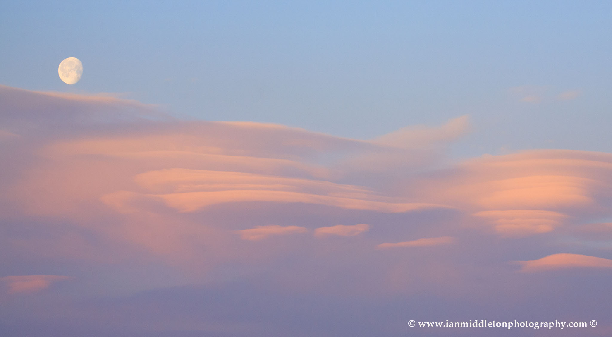Lenticular cloud formation (Altocumulus lenticularis) at sunrise with the waning moon setting above then over residential area of Ljubljana taken from a sixth floor apartment , Slovenia